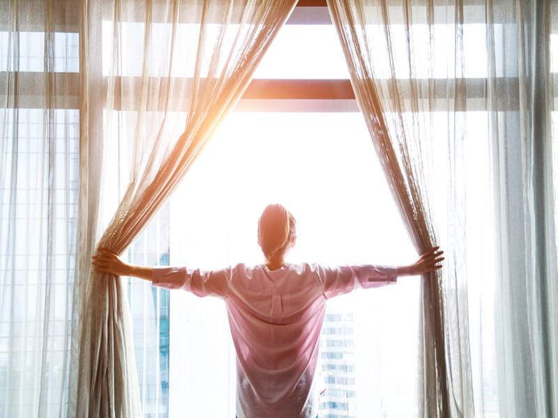 woman opens curtains to sunrise